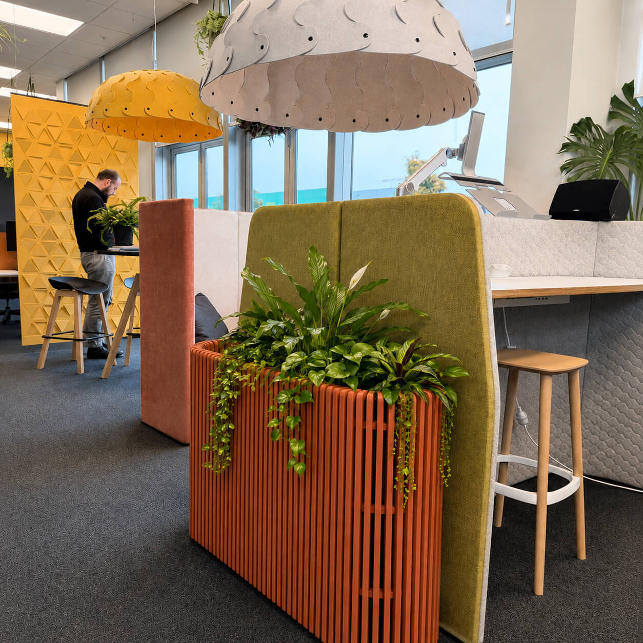 A modern office interior with a prominent orange slatted planter box holding green trailing plants, set against a white desk and gray carpet. A person is visible in the background near yellow geometric wall panels and pendant lights.