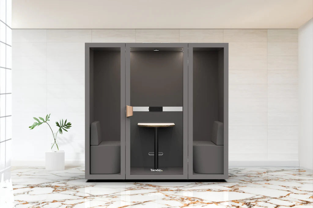 A modern meeting booth with clear glass walls and a fabric cushioned door, situated in a well-lit, open-plan office space. There are people seated and working in the background.